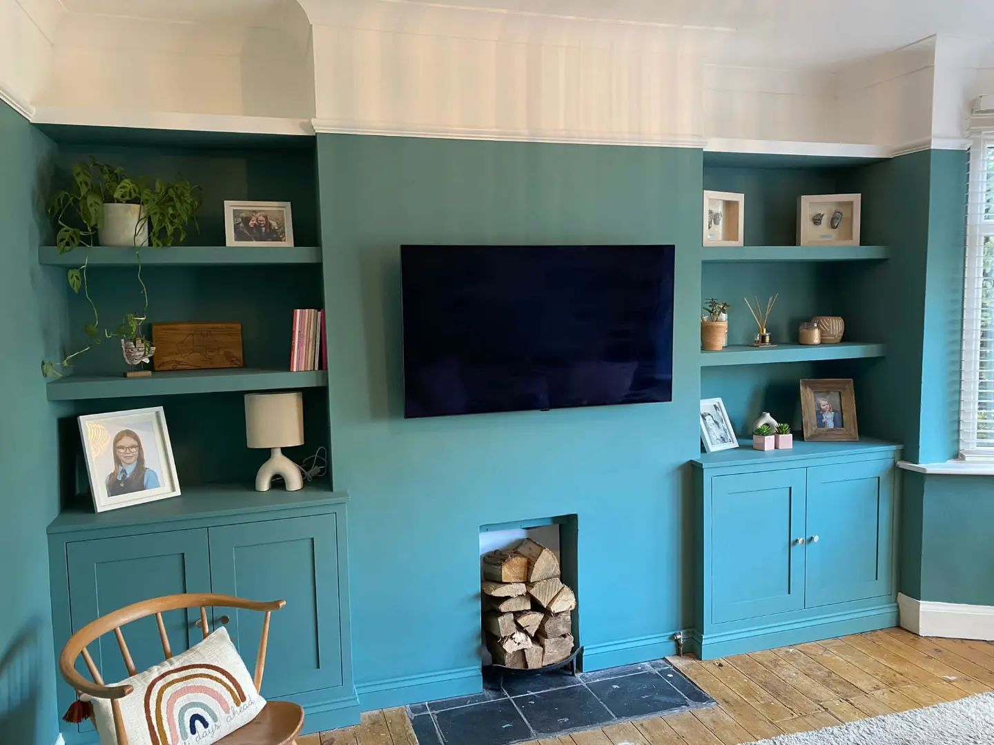 Teal-painted custom alcove cabinetry and floating shelves by a West Midlands joinery, framing a cozy fireplace stocked with firewood, with a wall-mounted TV above, and tastefully decorated with plants, photos, and books, adding a touch of personal style to a traditional UK home living room.