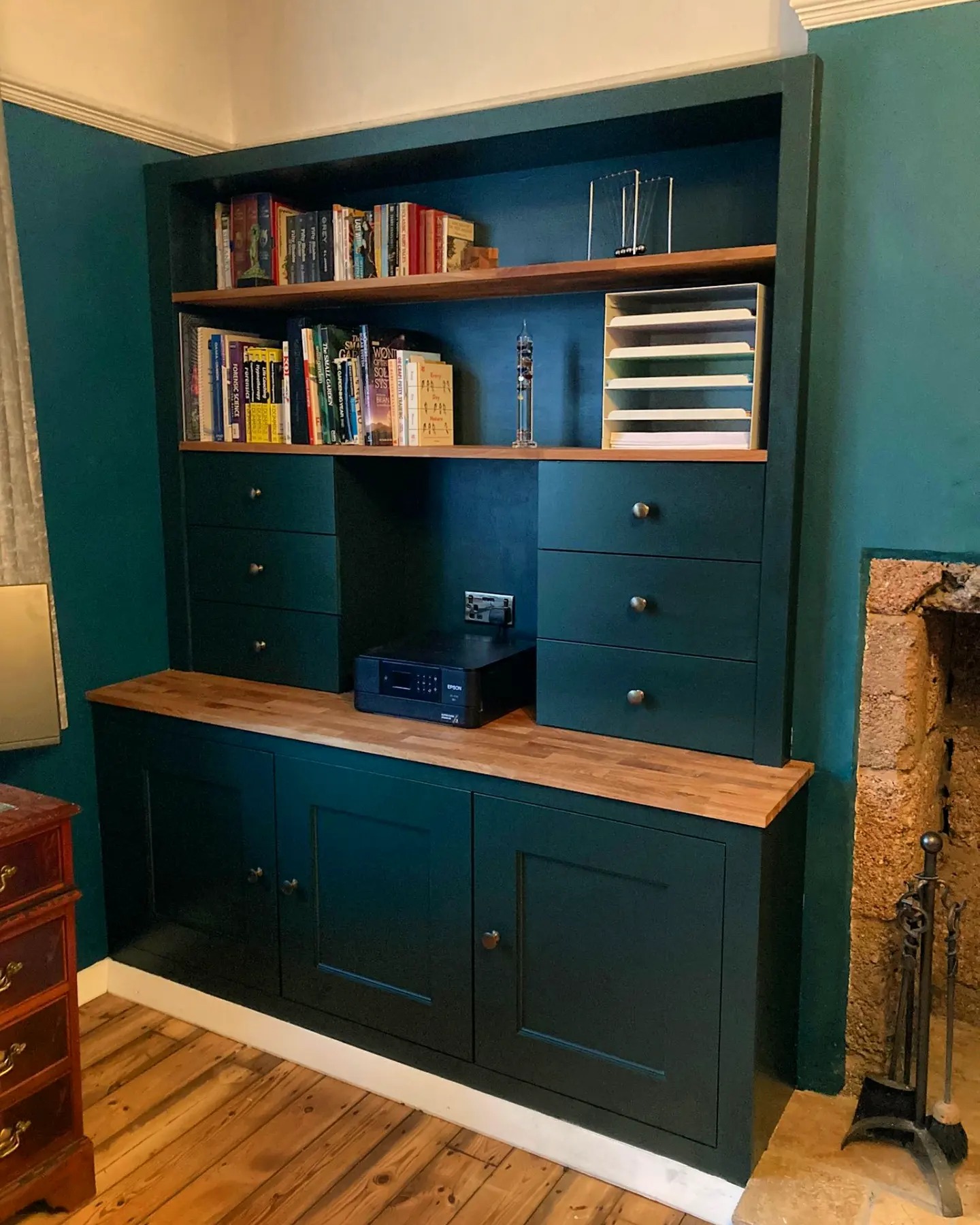 Stylish dark teal bespoke cabinetry with open shelving and wooden countertop, crafted by a West Midlands carpenter, offering a custom-built storage solution that harmoniously blends with the room's rustic charm and natural wood flooring, perfect for organizing books and displaying decor in a UK home.