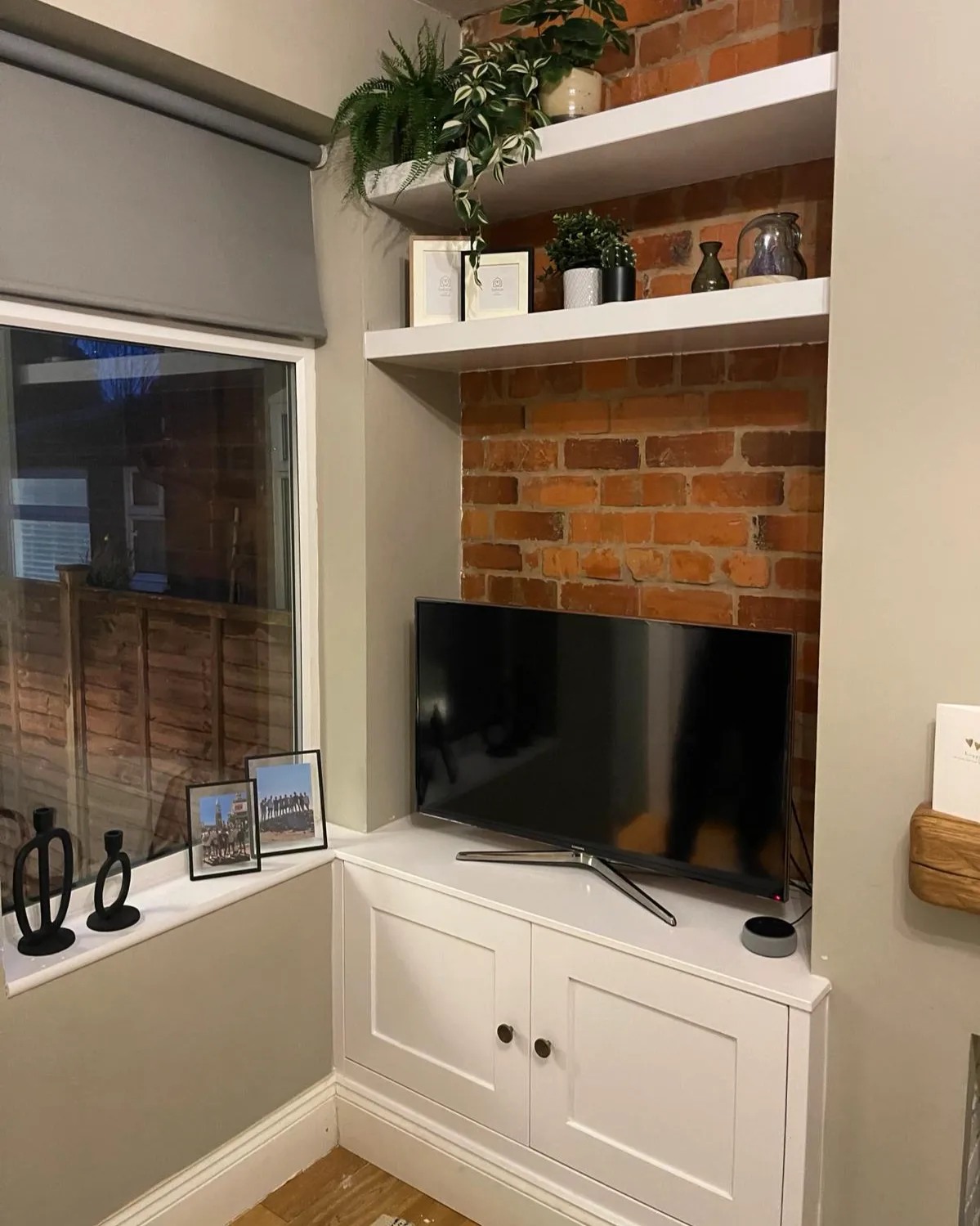 Contemporary living room corner featuring custom-built white cabinetry and floating shelves against an exposed brick wall by a West Midlands joiner, complemented with a flat-screen TV and tasteful greenery, creating a chic and functional space in a UK home with herringbone wood flooring.