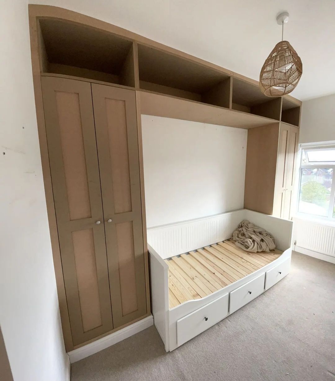 Custom-crafted fitted wardrobe with integrated bed and storage drawers, unfinished wood and MDF design, awaiting final touches by a local West Midlands joiner, featuring a contemporary aesthetic with ample shelving above and a cozy nook, illuminated by a stylish woven pendant light.