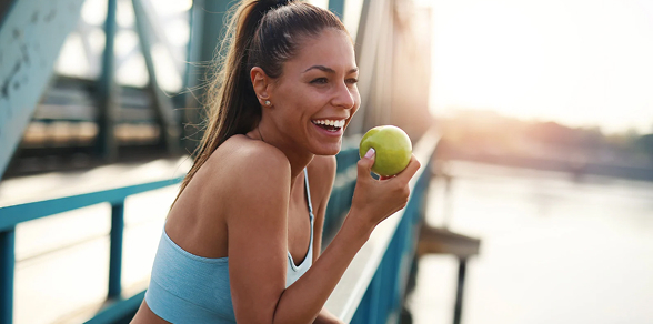 Fit young woman in workout gear smiling and holding a green apple outdoors, symbolizing health, energy, and wellness.