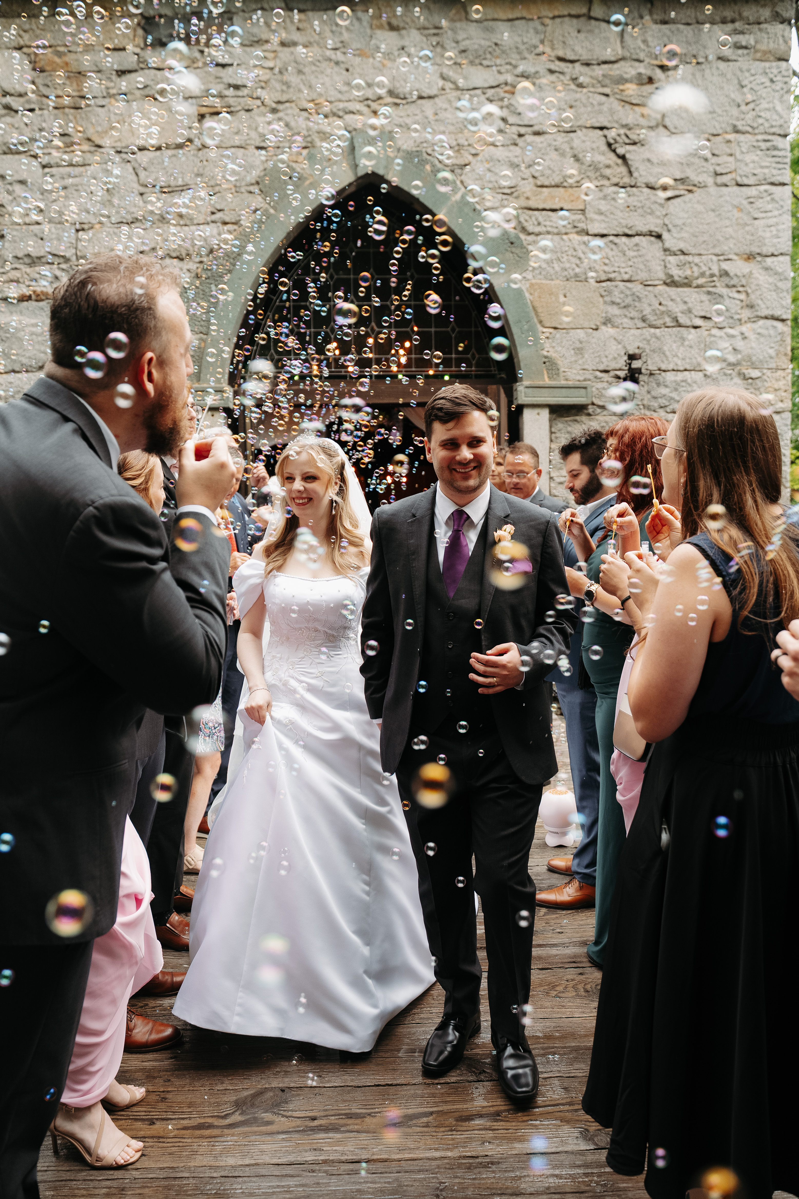 Couple walking out through a stone arch as guests blow bubbles