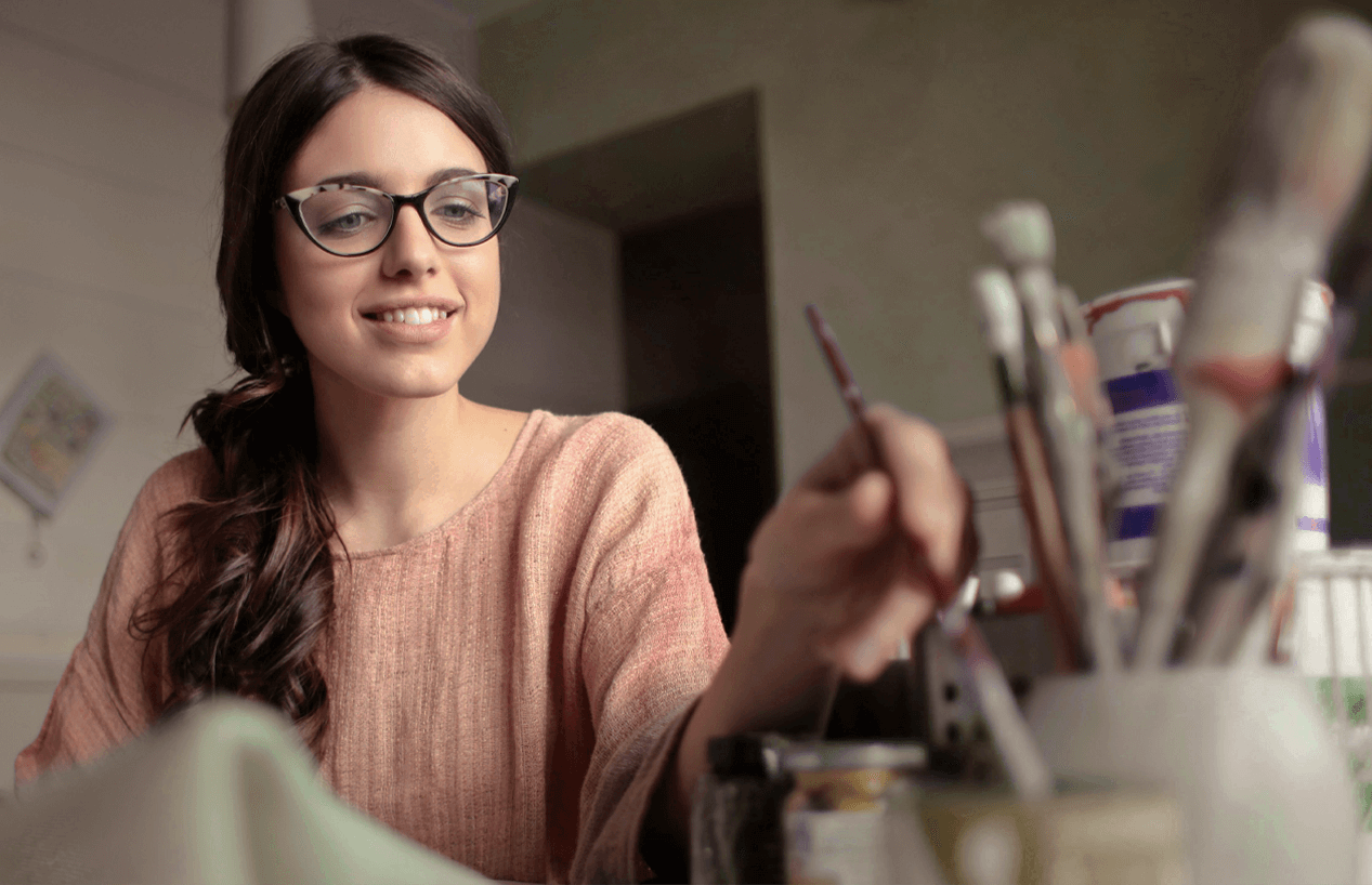 A smiling woman in a peach-colored sweater sits at a table, surrounded by art supplies, as she dips her paintbrush to add water to mix her pigments with.