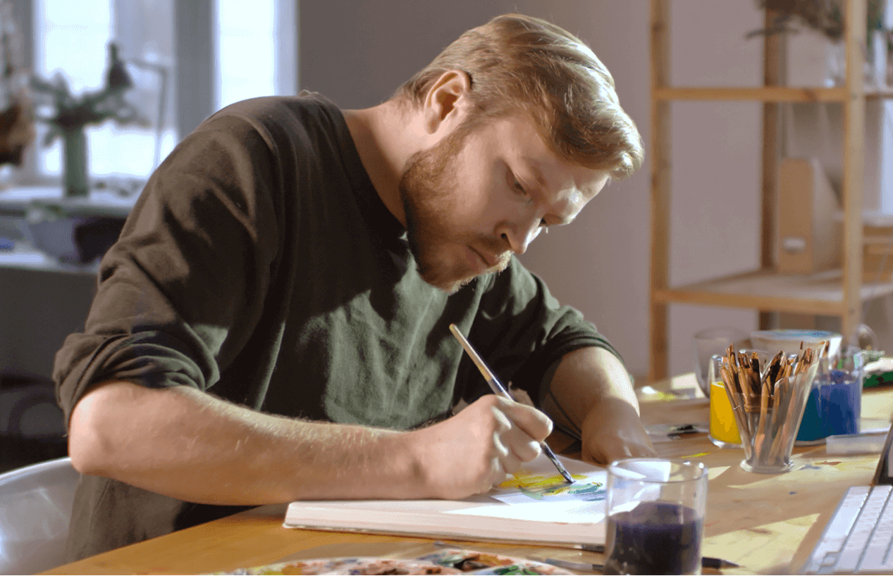 A man concentrating on his painting, surrounded by brushes and colorful paint in a well-lit studio.