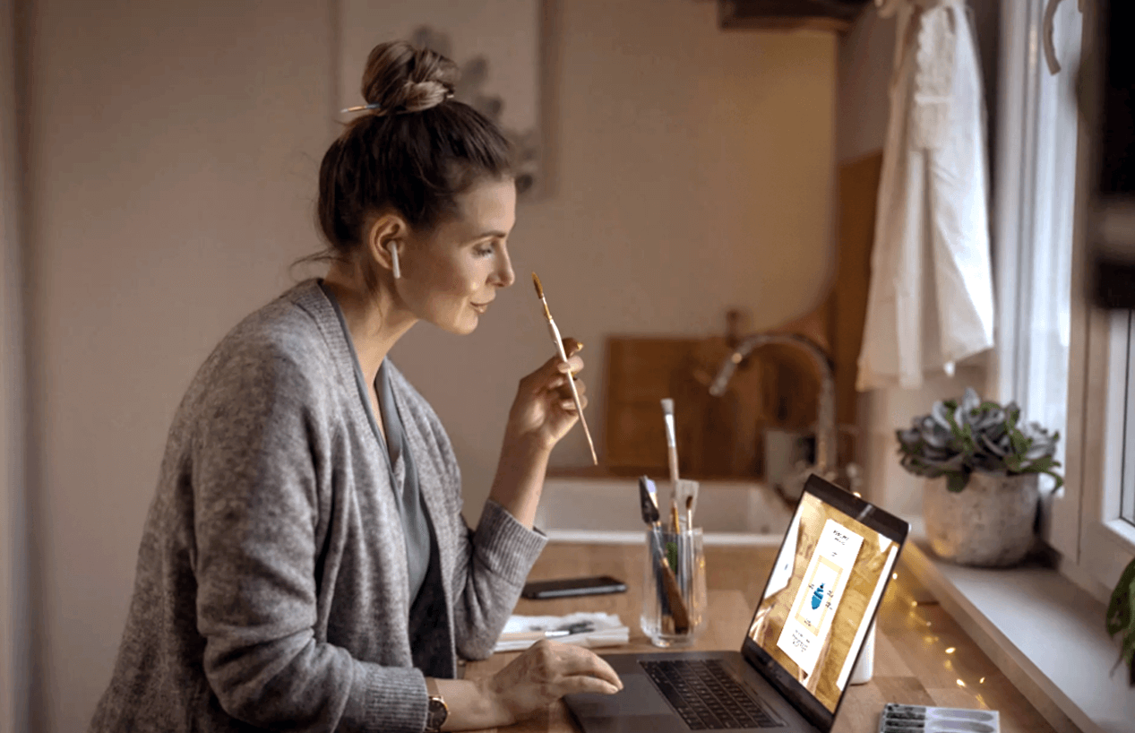 A woman smiling in a cozy setting while studying a painting process on a laptop, holding a brush thoughtfully. Art supplies and a plant are on the table.