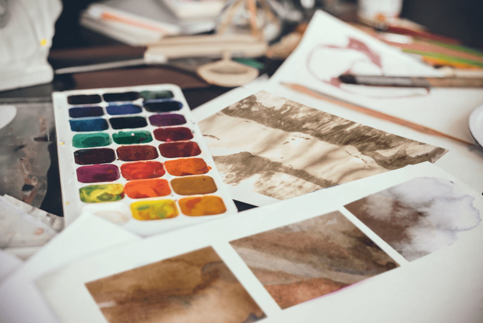 A smiling woman in a peach-colored sweater sits at a table, surrounded by art supplies, as she dips her paintbrush to add water to mix her pigments with.