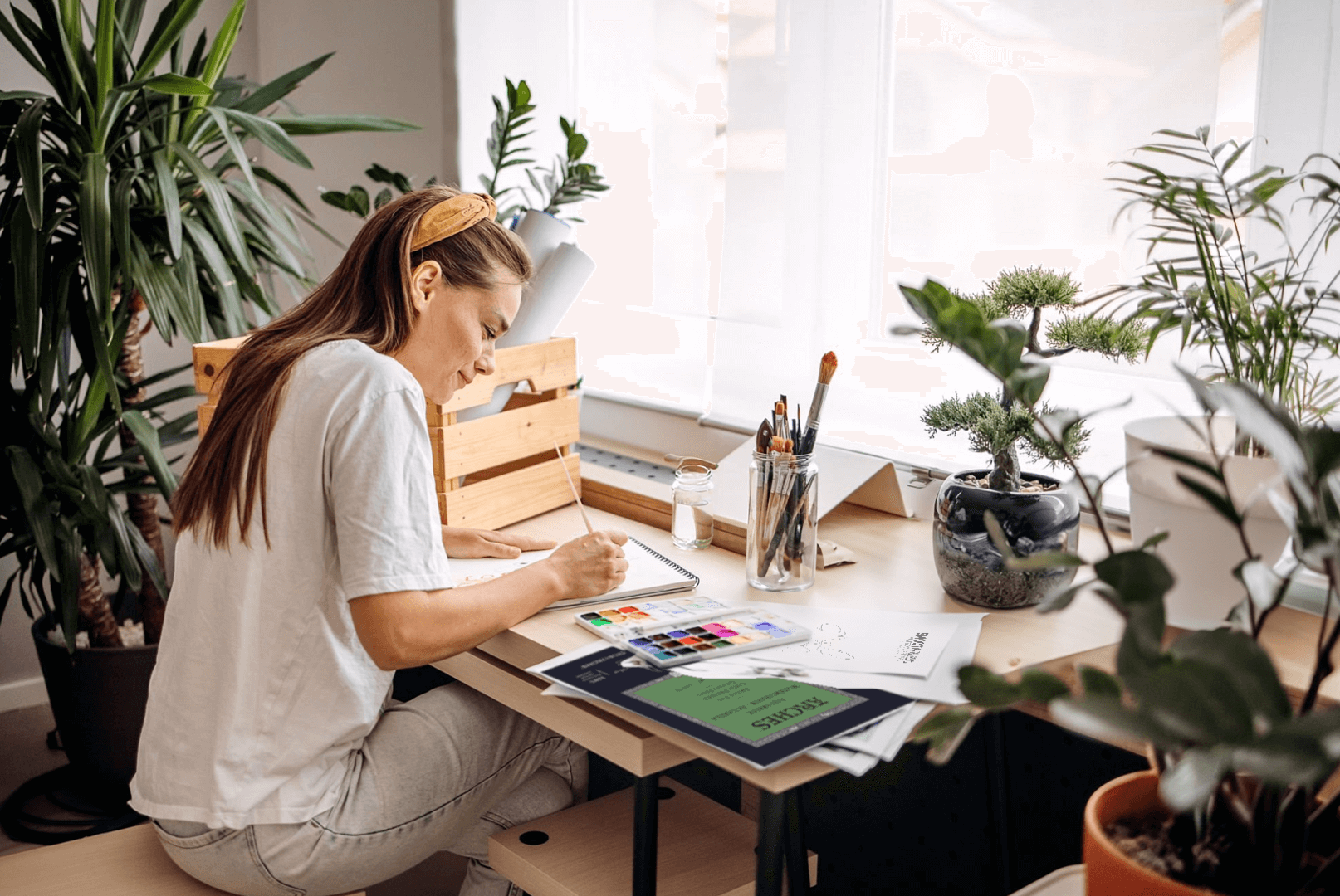 A woman smiling in a cozy setting while studying a painting process on a laptop, holding a brush thoughtfully. Art supplies and a plant are on the table.