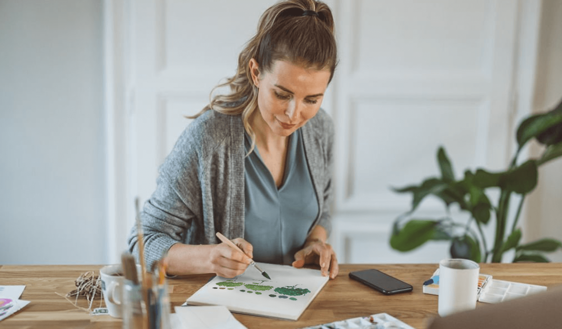 A smiling woman in a peach-colored sweater sits at a table, surrounded by art supplies, as she dips her paintbrush to add water to mix her pigments with.