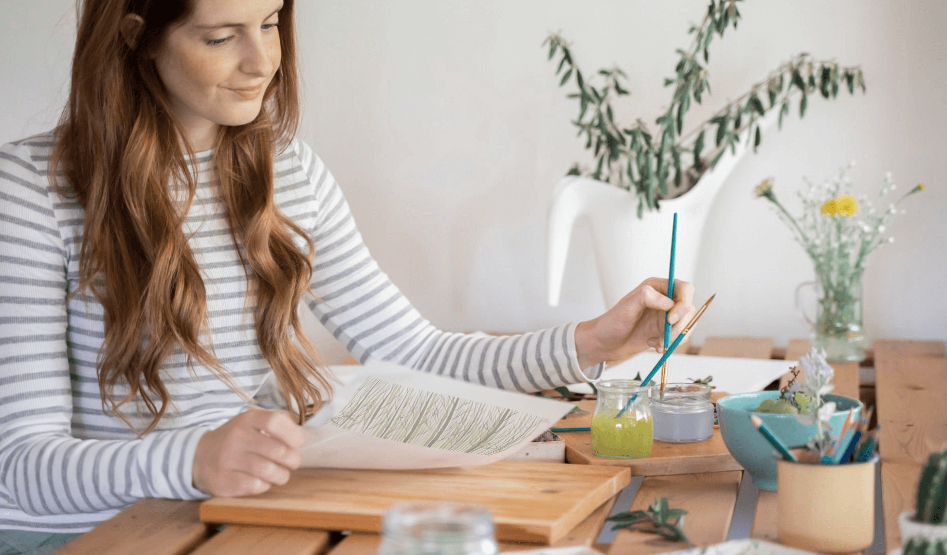 A smiling woman in a peach-colored sweater sits at a table, surrounded by art supplies, as she dips her paintbrush to add water to mix her pigments with.