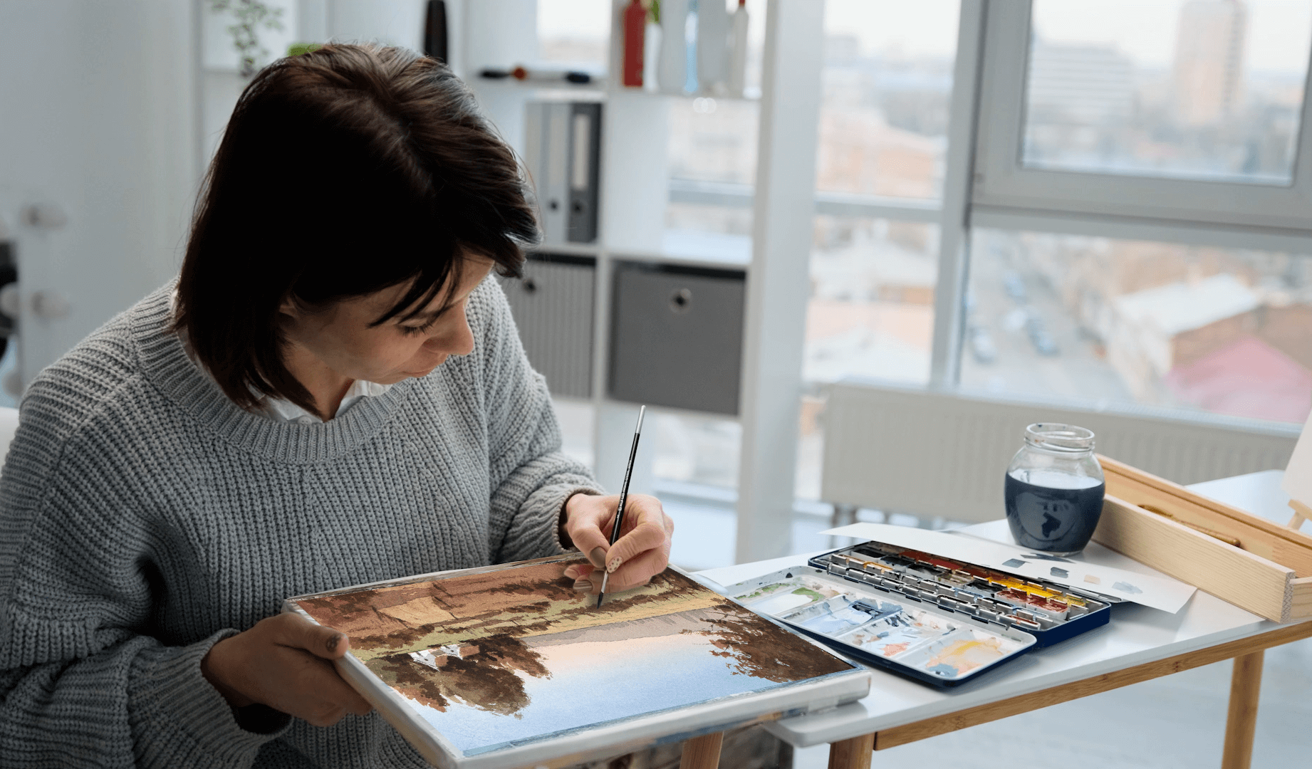 A smiling woman in a peach-colored sweater sits at a table, surrounded by art supplies, as she dips her paintbrush to add water to mix her pigments with.