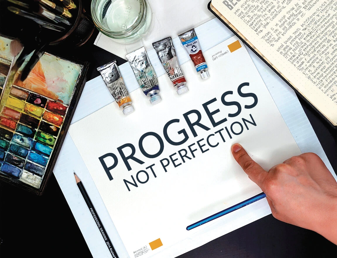 Hand pointing to a “Progress Not Perfection” note among watercolor supplies.