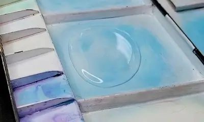 A watercolor palette with shades of blue and purple features a dome-shaped water droplet at the center, reflecting light. The scene is calm and artistic.