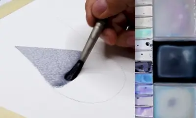 A hand paints a grey triangle on paper using a brush. A watercolor palette with various shades is visible to the right, indicating a calm, creative process.