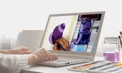 A person paints a purple seashell on a laptop screen, with paints and brushes nearby. The scene conveys creativity and digital art creation.