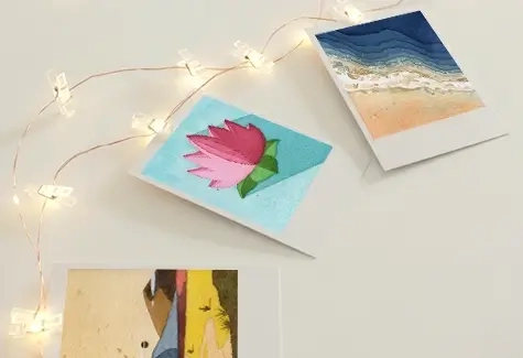 Three art prints are spread on a white surface, featuring a pink flower, a beach scene, and an abstract design. String lights add a warm glow.