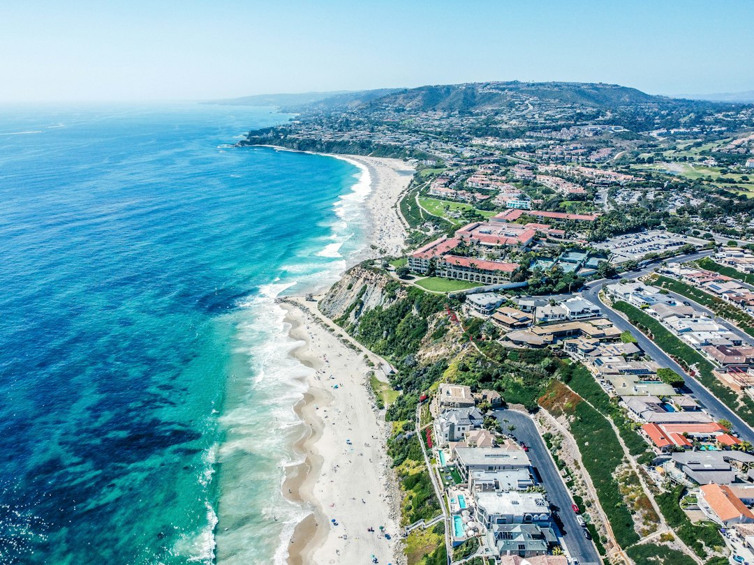 Aerial view of a Southern California shoreline with turquoise water, wide beach and hillside homes and resorts.