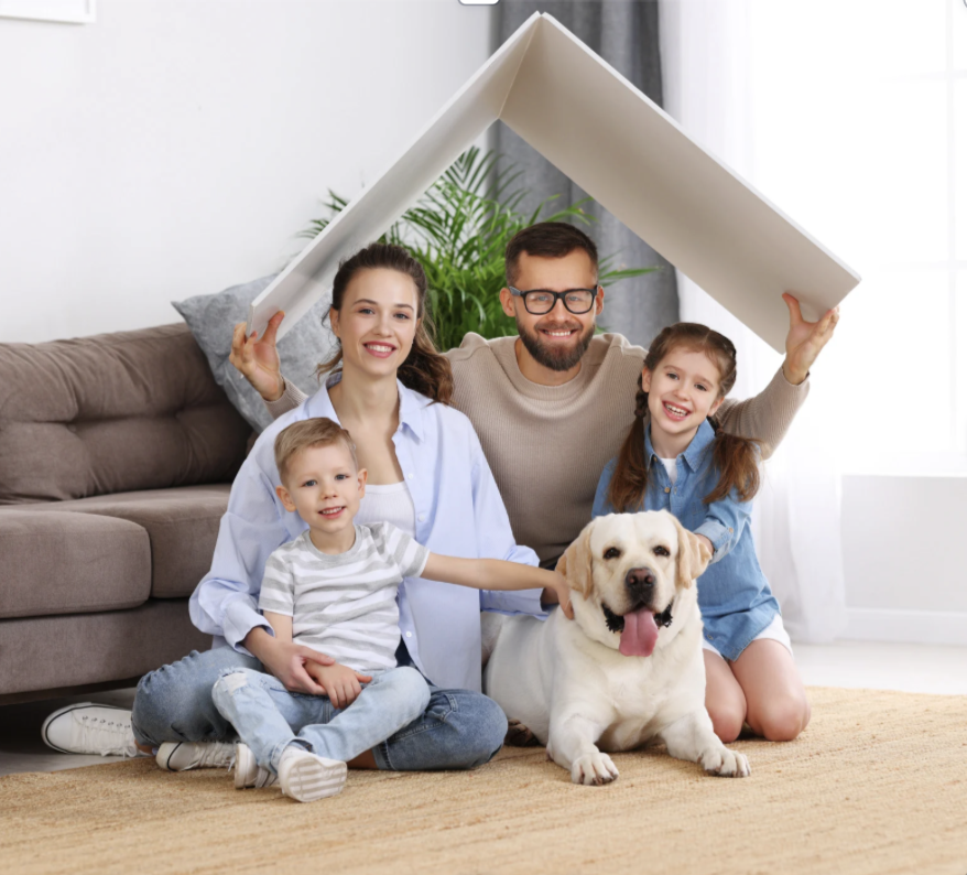Happy family with two children and a Labrador dog sitting on a living room floor, holding a roof-shaped board above their heads to symbolize homeownership, in a bright modern home interior.