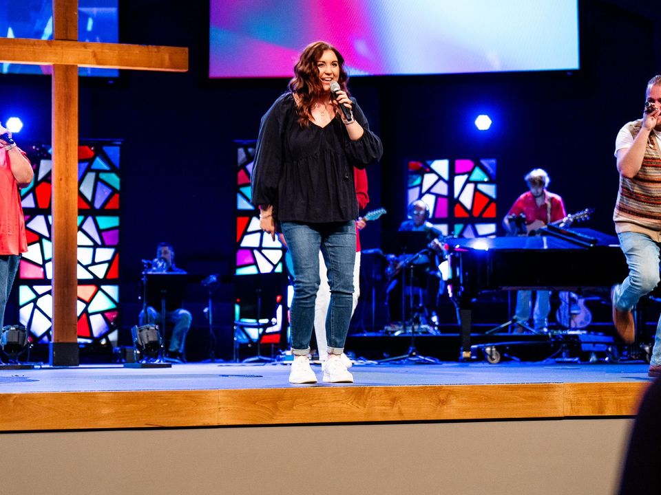Church stage with a large wooden cross, colorful stained-glass panels and a band, with a singer at center holding a microphone