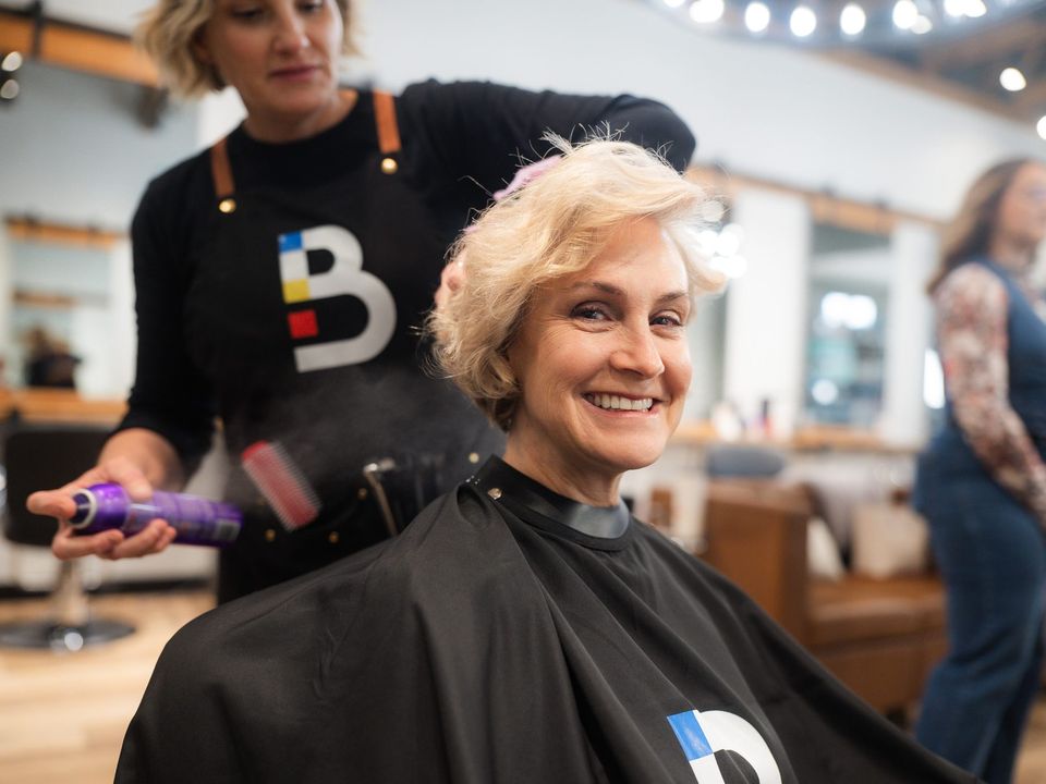 Stylist in a black branded apron spraying product as she styles a smiling client's hair in a salon chair