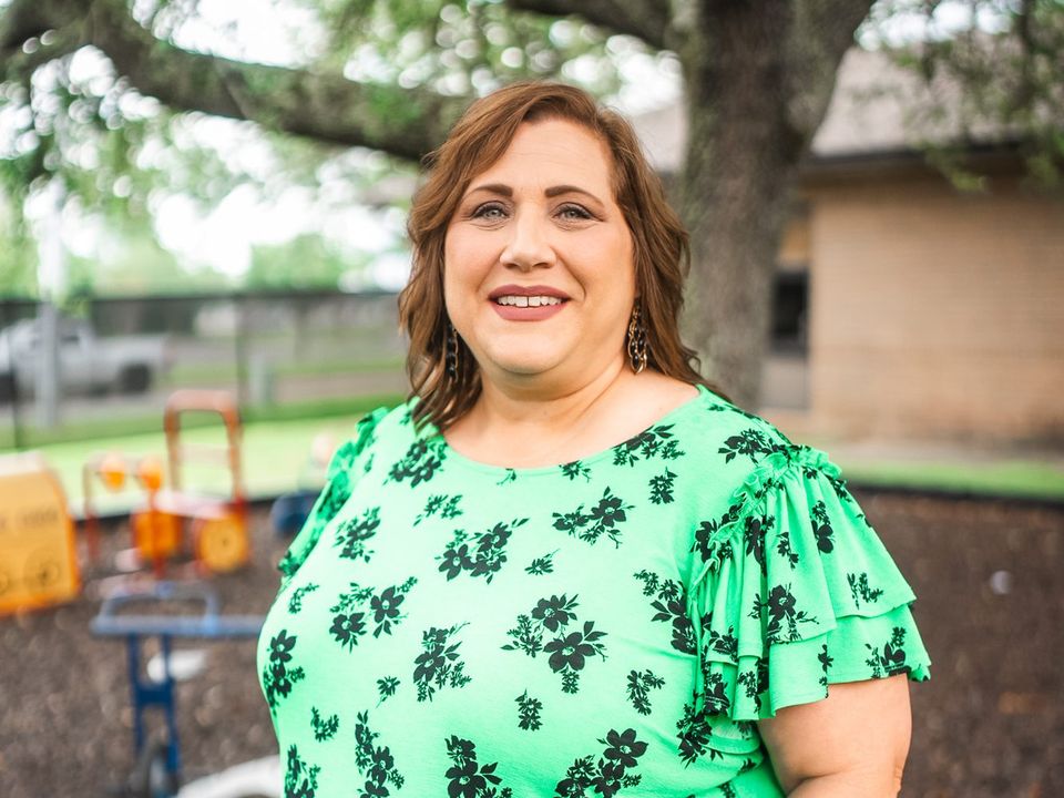 Smiling woman in a green floral top standing under oak trees, with a preschool playground blurred behind her