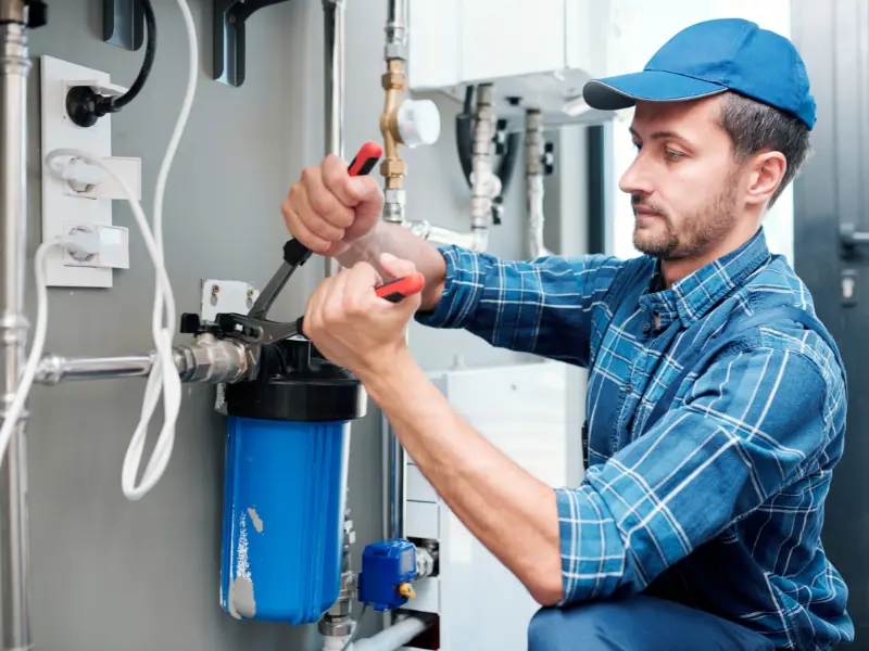 Plumber installing and adjusting a whole-house water filter system on a mechanical room wall.