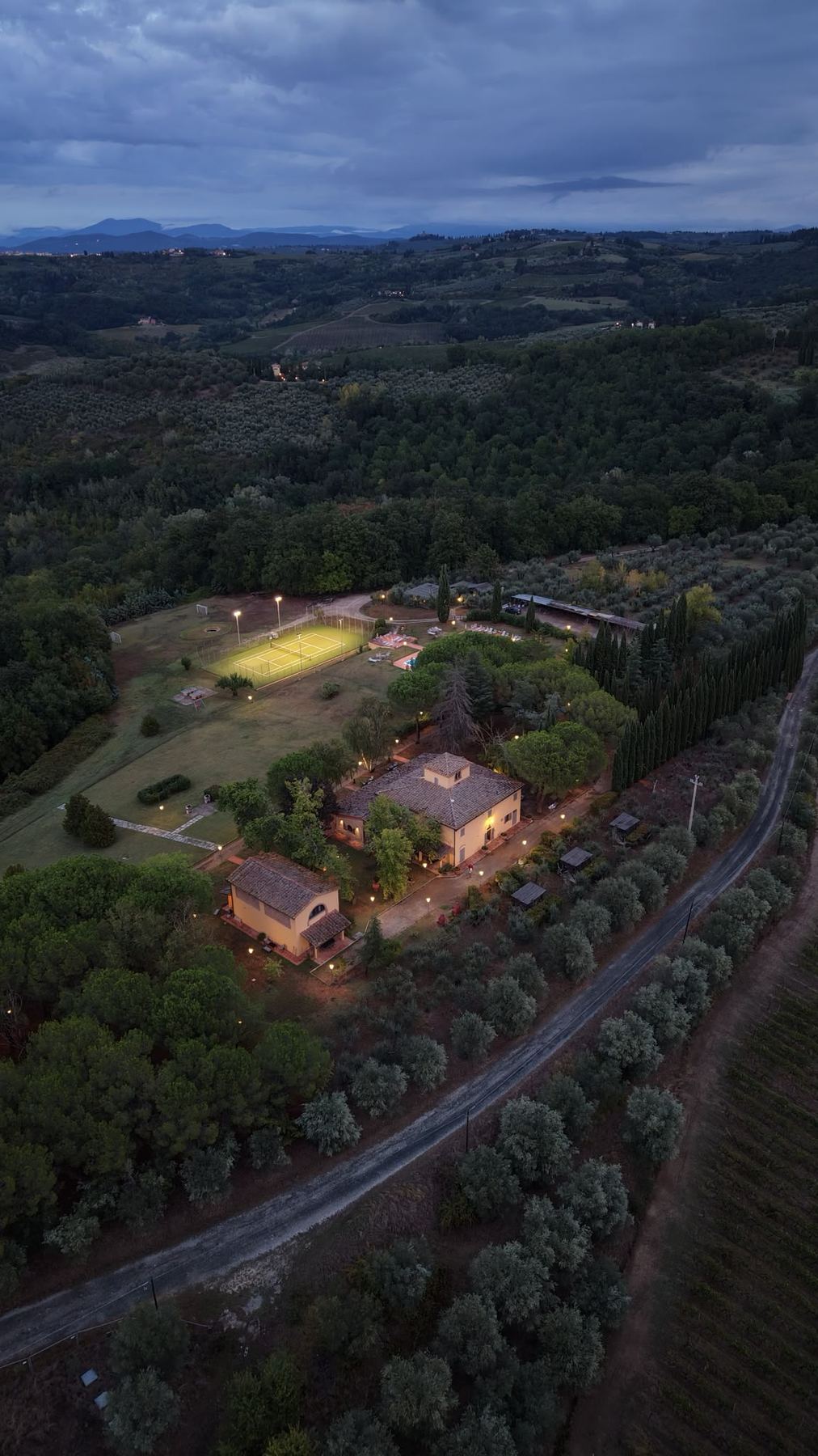 The estate at dusk from the air, lamps glowing along the paths and the tennis court lit up
