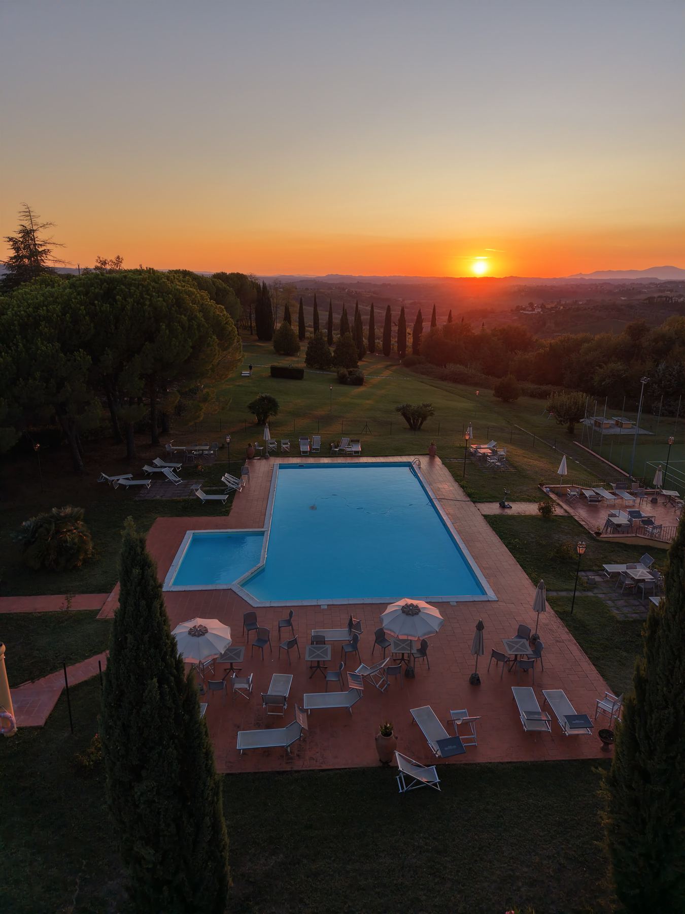 Sunset over the Tuscan hills seen from above the pool terrace, with cypress trees and umbrellas