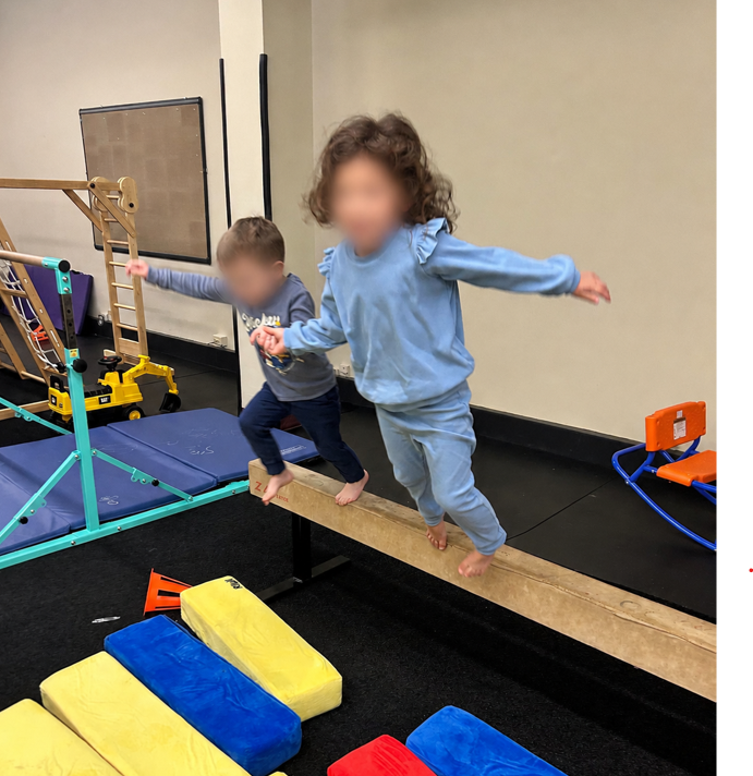 Two young children balancing barefoot on a wooden beam during an indoor movement activity.