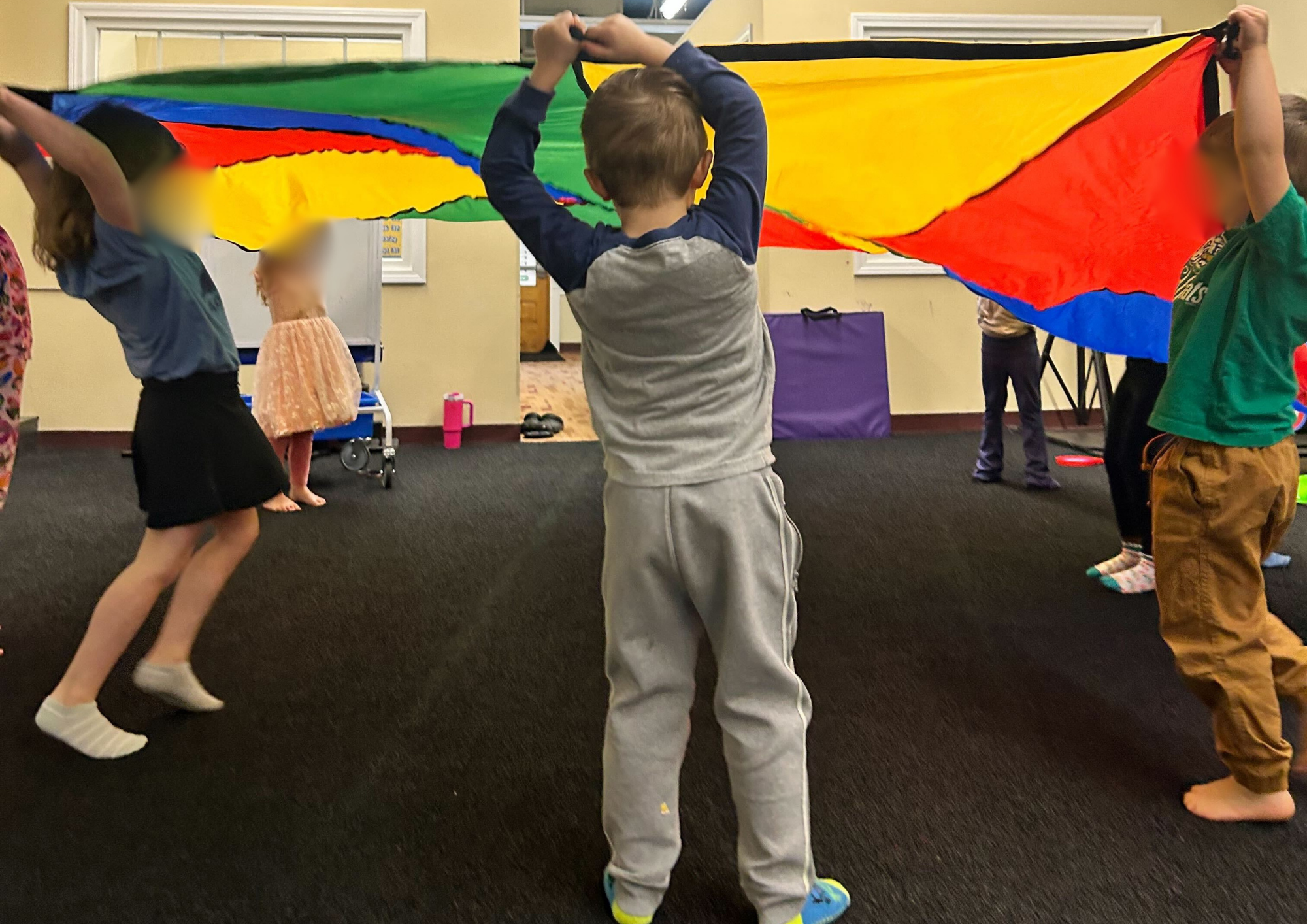 Children engaging in active group play indoors while holding a colorful parachute overhead, encouraging movement, coordination, teamwork, and social interaction.