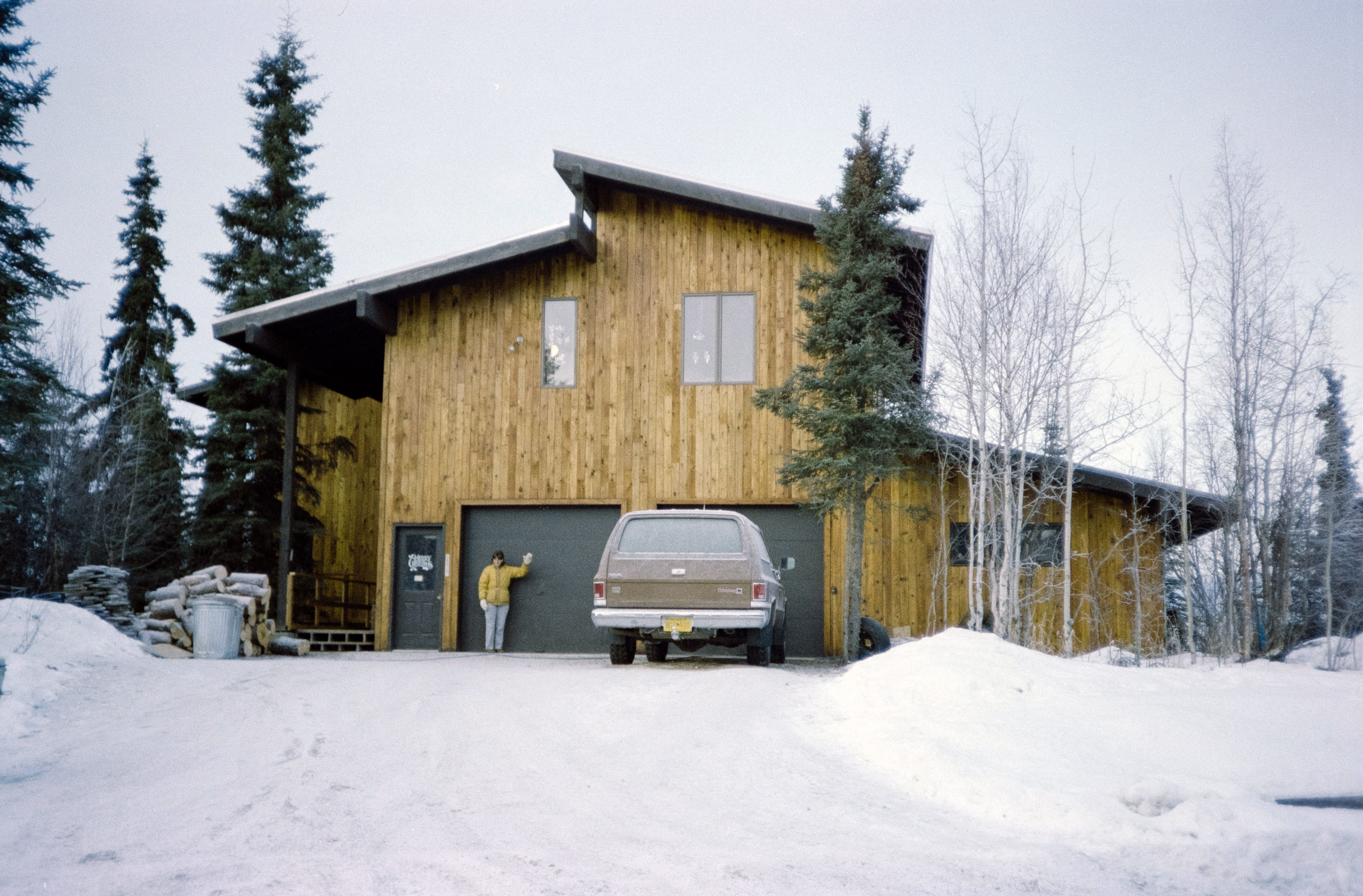 A snow-covered wooden home in Alaska, built by Skye’s father, surrounded by evergreen and bare trees, with a truck parked outside and a person standing near the garage.
