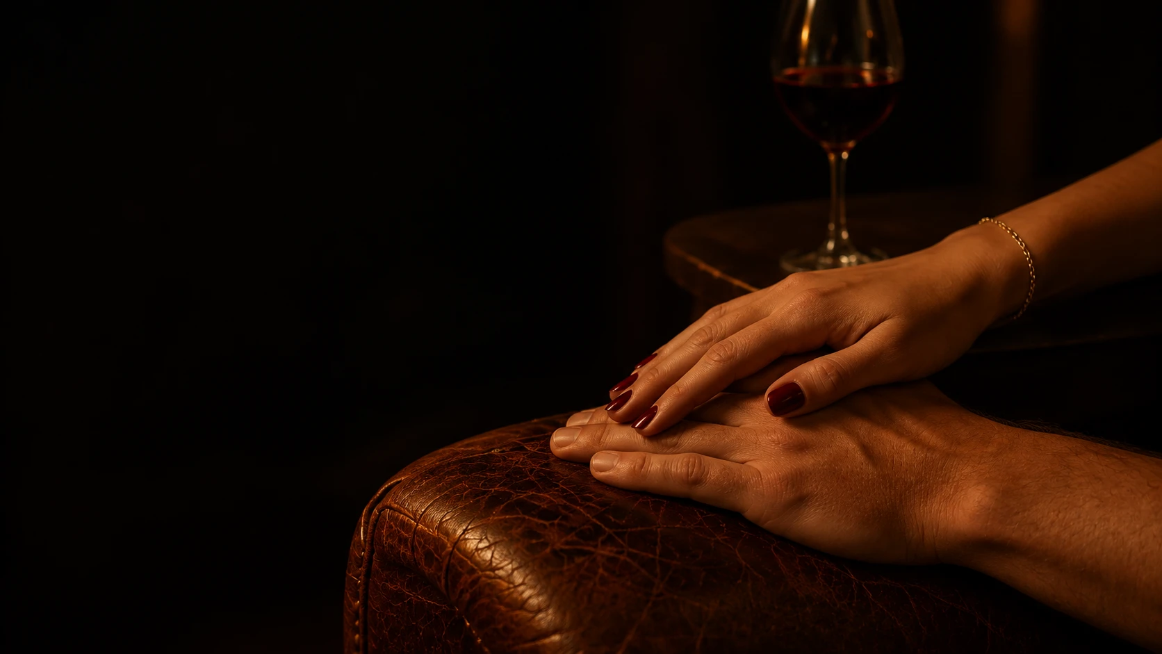 Dos manos se rozan sobre un sillón de cuero, junto a una copa de vino.