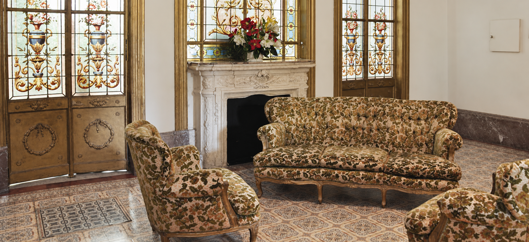 Salón con sillones de época, chimenea de mármol y vitrales florales.