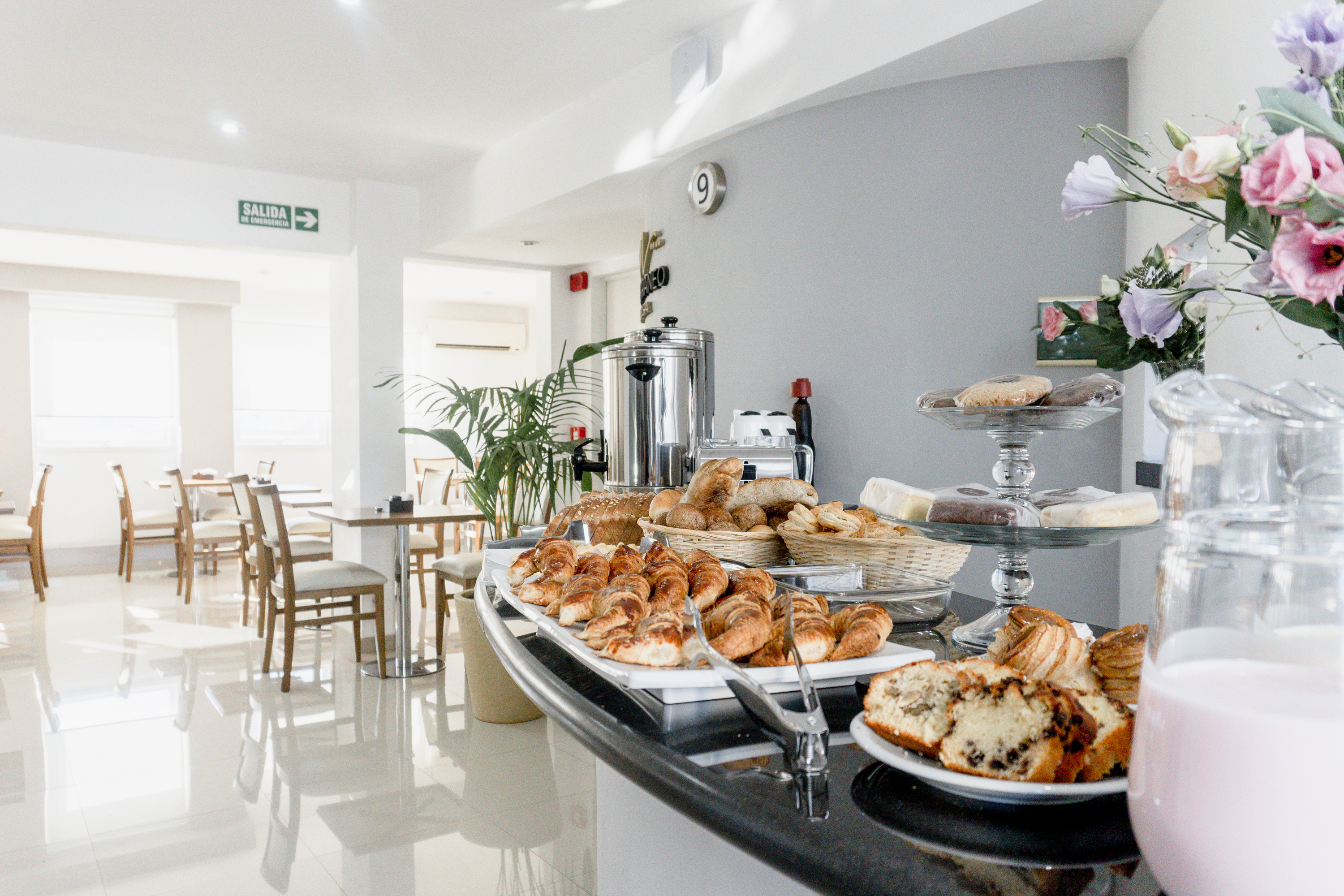 Desayuno buffet con medialunas, panes, tortas y bebidas en un salón luminoso.