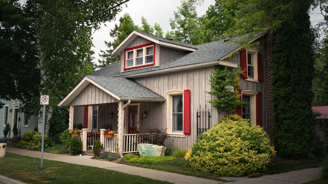 Grey-sided bungalow with red window trim, covered front porch, flowering shrubs and a mown lawn on a quiet residential street.