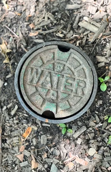 Close-up of a metal water access cover embedded in the ground, surrounded by dirt and mulch, used to control or monitor underground water lines.