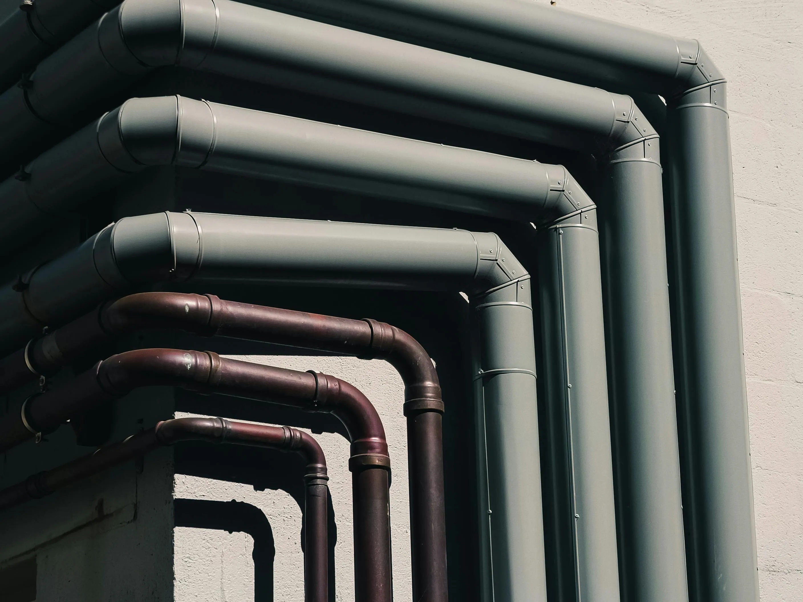 Multiple industrial pipes of varying sizes and materials running along the side of a building, illustrating complex plumbing infrastructure.