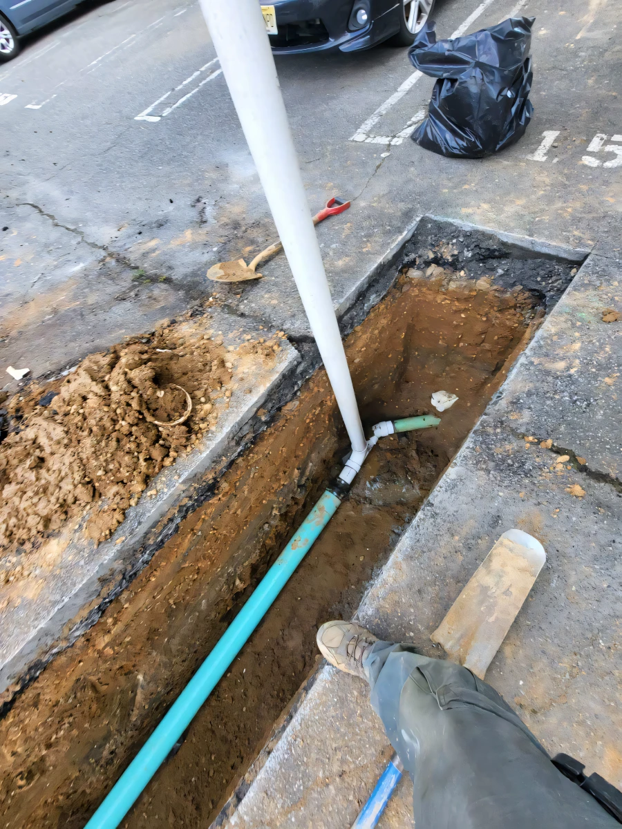 Worker standing near an open trench with exposed green pipes and tools on a paved area