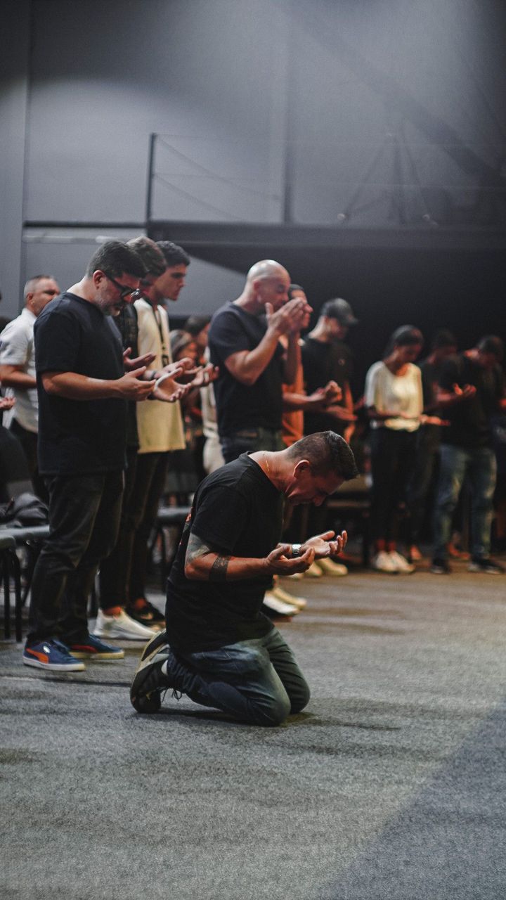 Man kneeling in prayer during a Christian worship service while others stand with hands lifted in devotion, representing heartfelt worship, faith, repentance, and spiritual surrender in church.