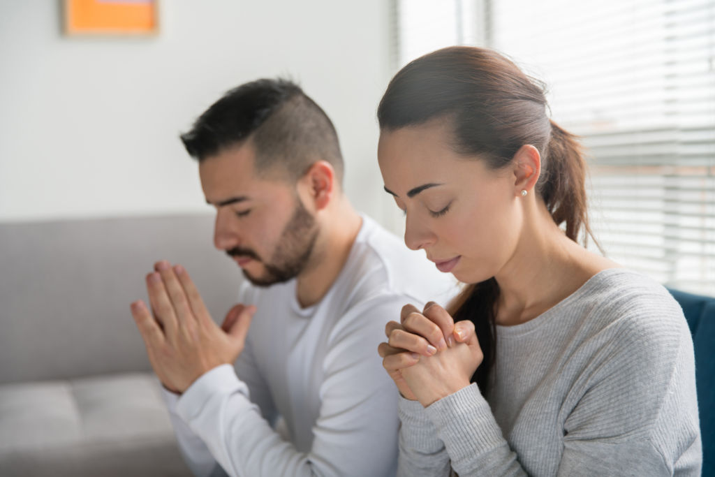 Young Christian couple praying together at home with eyes closed and hands clasped, symbolizing spiritual intimacy, unity, and faith in marriage. Young Christian couple praying together at home with eyes closed and hands clasped, symbolizing spiritual intimacy, unity, and faith in marriage.