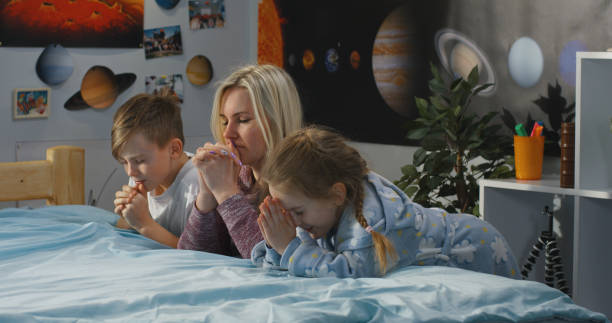 Mother and two young children kneeling in prayer beside a bed, demonstrating family prayer, Christian parenting, faith-filled home life, and spiritual growth through daily devotion.