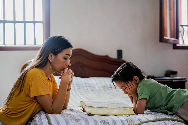 A godly mother and her young son lying on a bed with an open Bible, praying together with clasped hands — illustrating spiritual leadership for mothers and family discipleship in a faith-filled home.