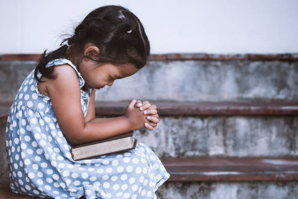 Young Christian child praying with a Bible while sitting on steps, learning to trust God and pray for others through faith, compassion, and biblical guidance.