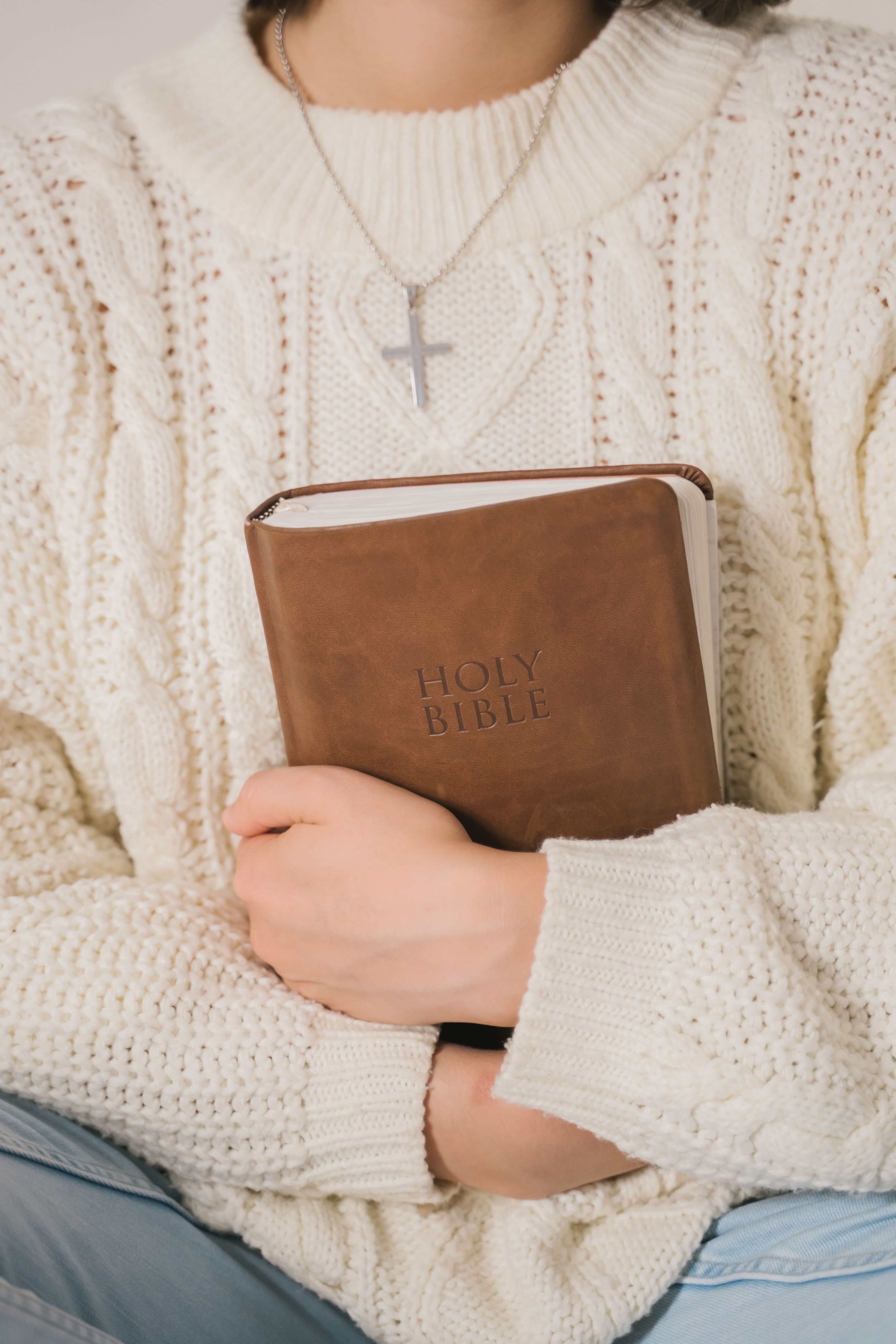 Christian mother holding a Holy Bible while wearing a white knitted sweater and cross necklace, representing faith, prayer, biblical parenting, and teaching children about God's love.