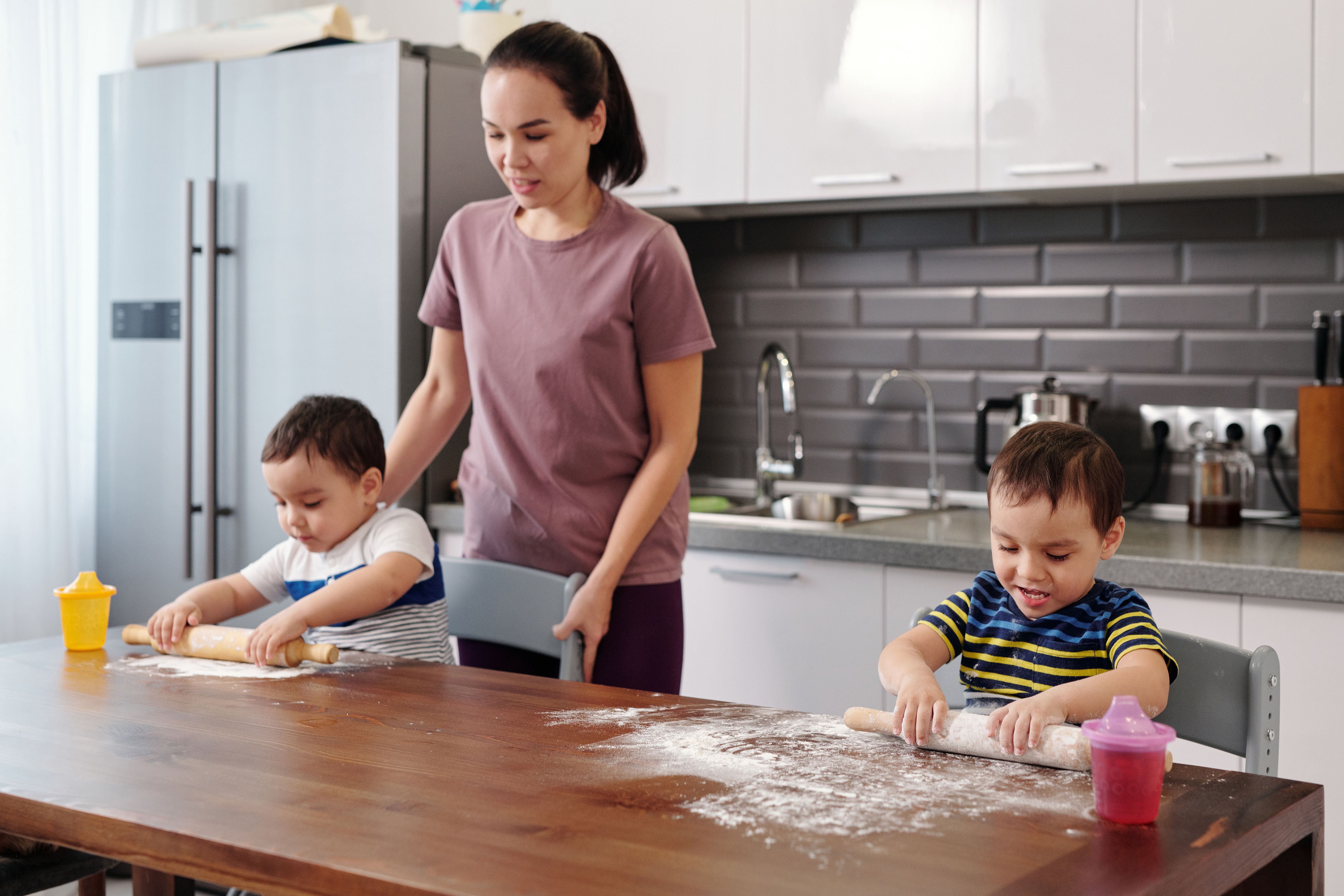 A mother teaching her young children basic cooking skills at home as they roll dough on a kitchen table, helping them develop practical life skills, independence, and confidence through family cooking activities. A mother teaching her young children basic cooking skills at home as they roll dough on a kitchen table, helping them develop practical life skills, independence, and confidence through family cooking activities.
