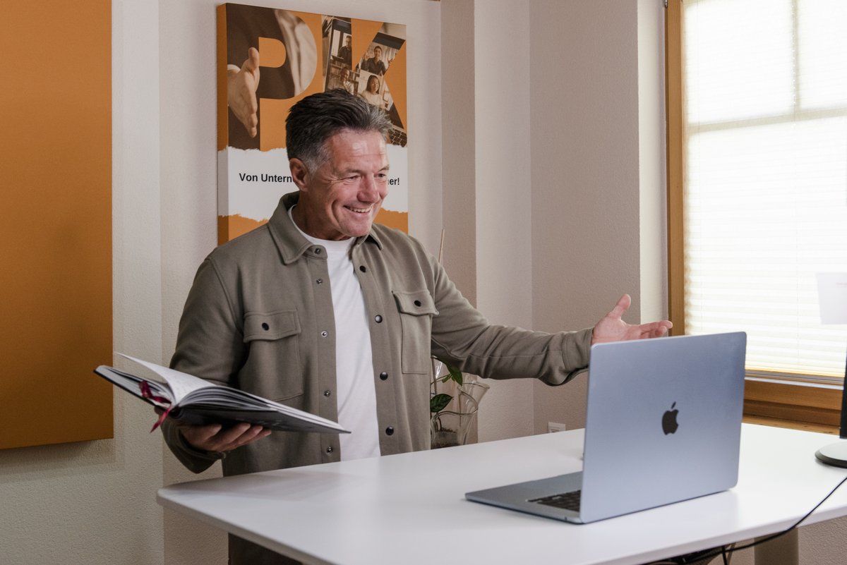 Peter Klenner am Schreibtisch, mit offenem Notizbuch, lacht und begr&uuml;&szlig;t die Runde am Laptop