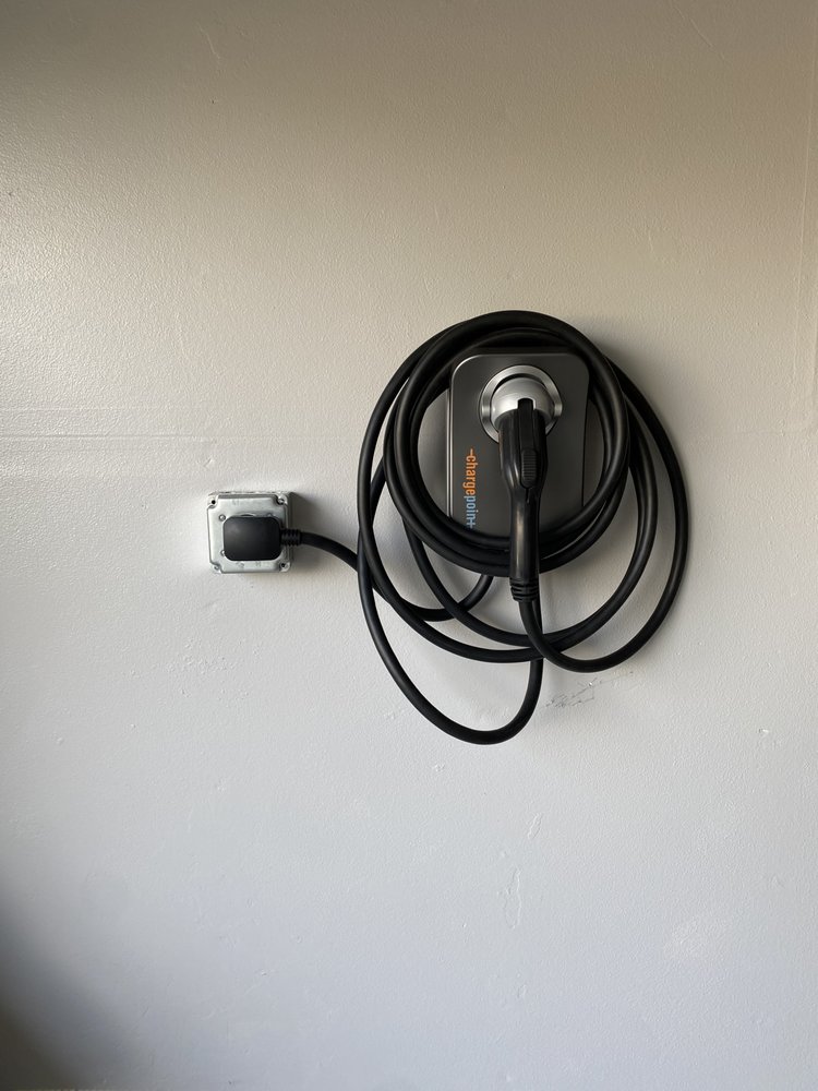 A Level 2 EV charger mounted on the interior wall of a residential garage in Orange County, CA, with clean conduit running to a nearby electrical panel. A white electric vehicle is parked in the garage with the charging cable connected.
