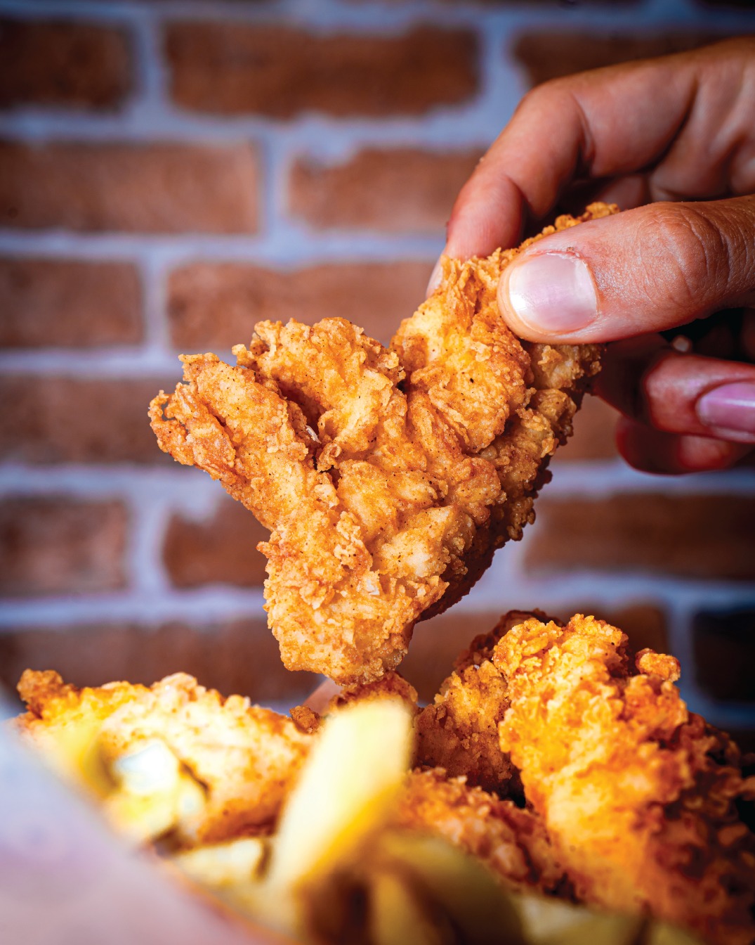 Hand grabbing a crispy fried chicken tender over a tray of tenders and fries