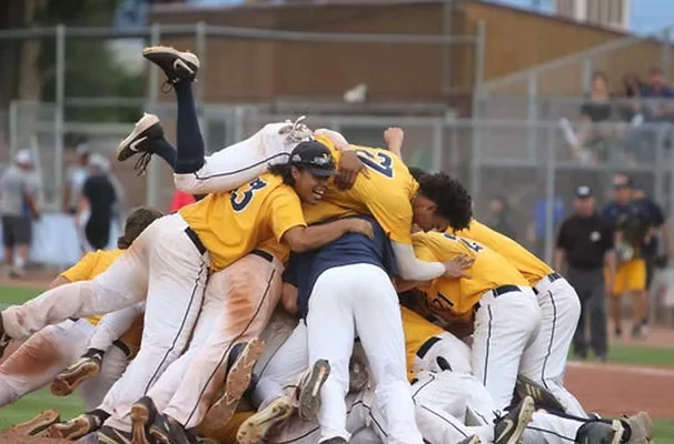 A baseball team celebrates victory in a dogpile on the field, with players in yellow and white uniforms.