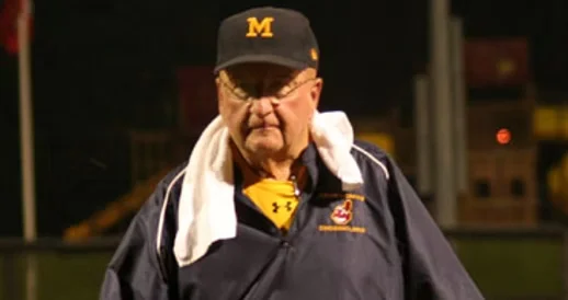 An older man wearing a black Michigan hat, glasses, and a navy jacket with a white towel over his shoulder, standing outdoors at night.