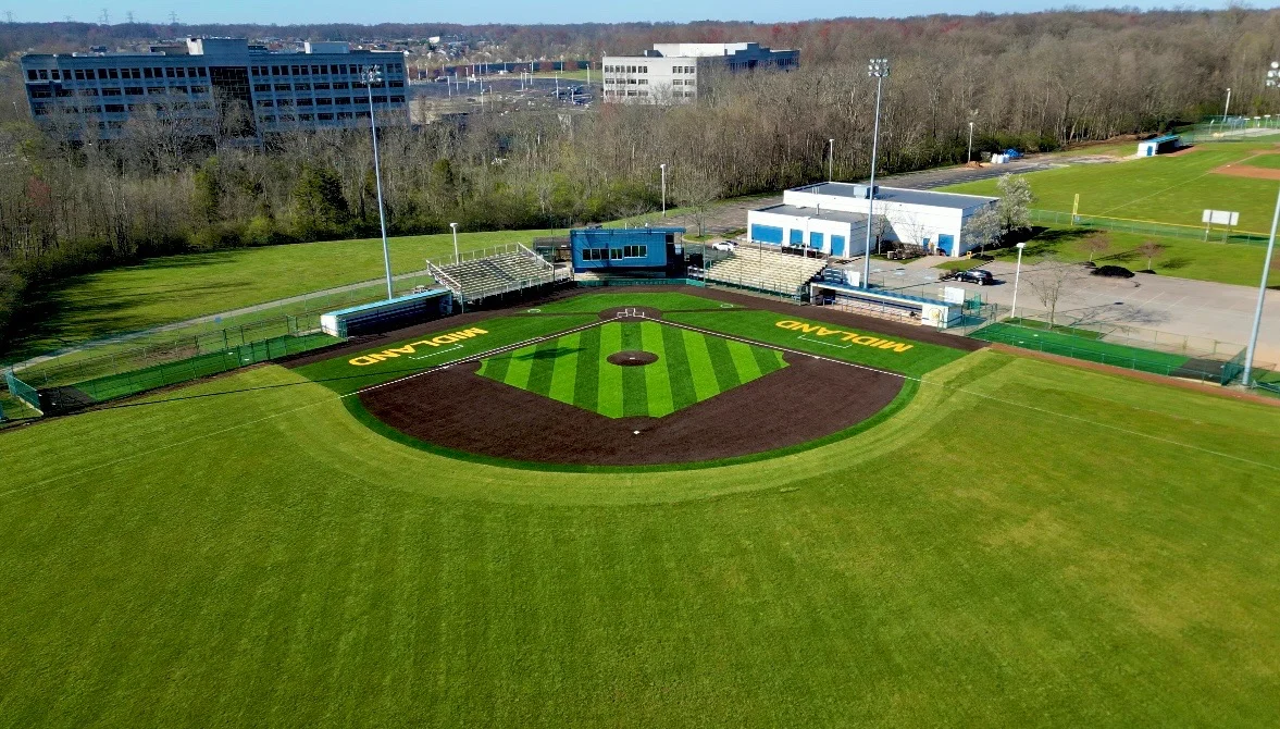 An aerial view of a vibrant green baseball field with 'MIDLAND' written on the outfield, surrounded by trees and buildings.